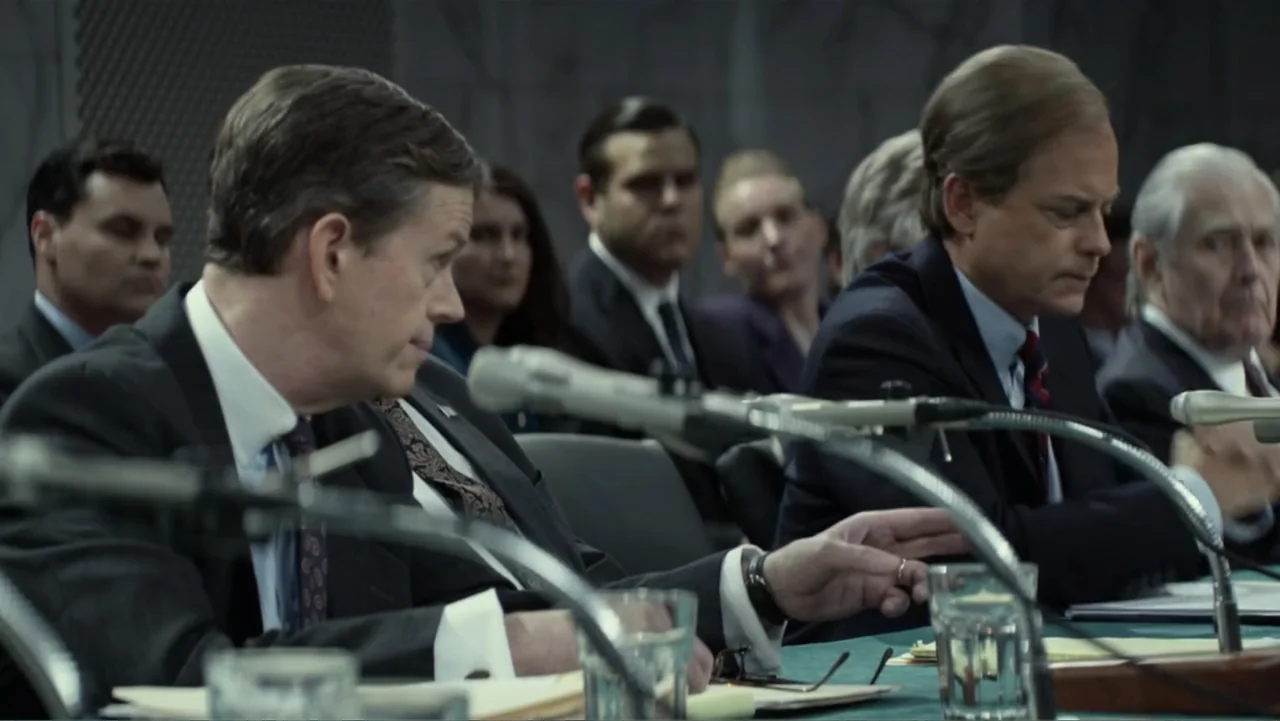 Confirmation (2016) movie still: medium — A formal hearing room features rows of desks and microphones at a table. Two men in suits sit at the front…