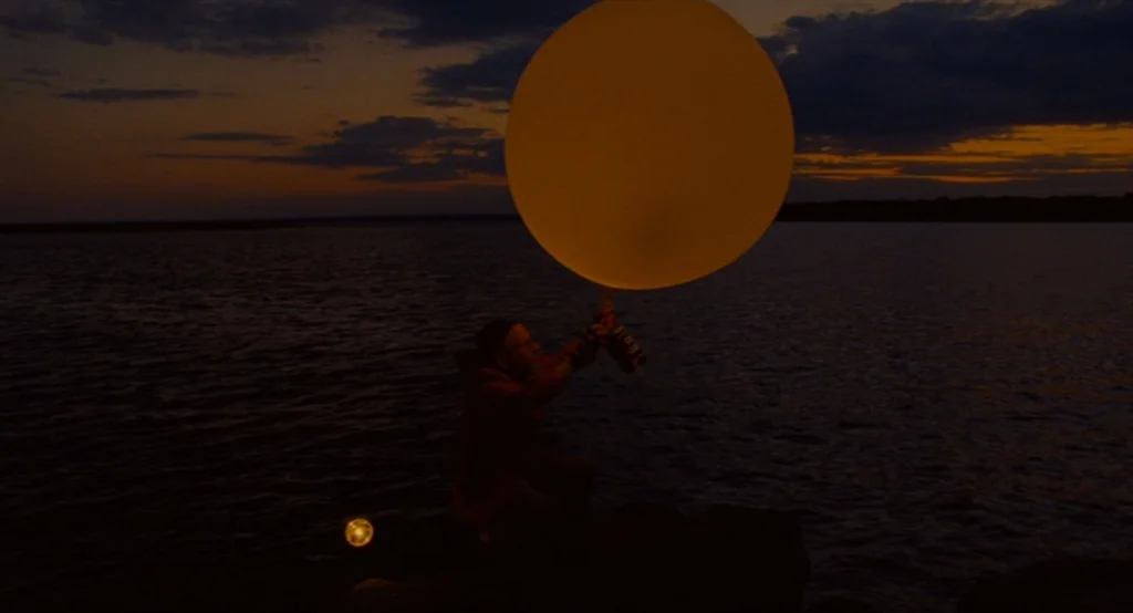 Moonrise Kingdom (2012) movie still: wide — A dark rocky shoreline sits beside a wide body of water at dusk. A person in a hooded jacket holds a…