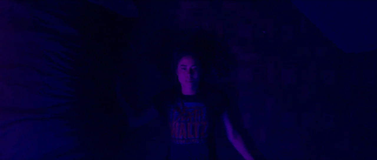 Bliss movie still: medium — A person lies on a dark, textured surface in a dimly lit room. Intense colored light casts purple and blue shadows over…
