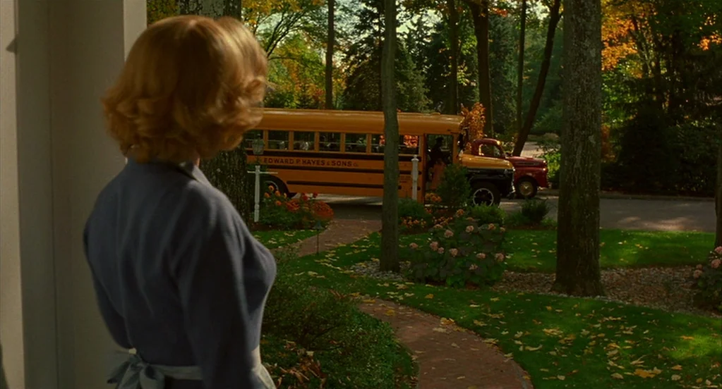 Far From Heaven (2002) movie still: medium — A suburban front yard features a winding brick path leading toward a school bus. A woman in a blue…