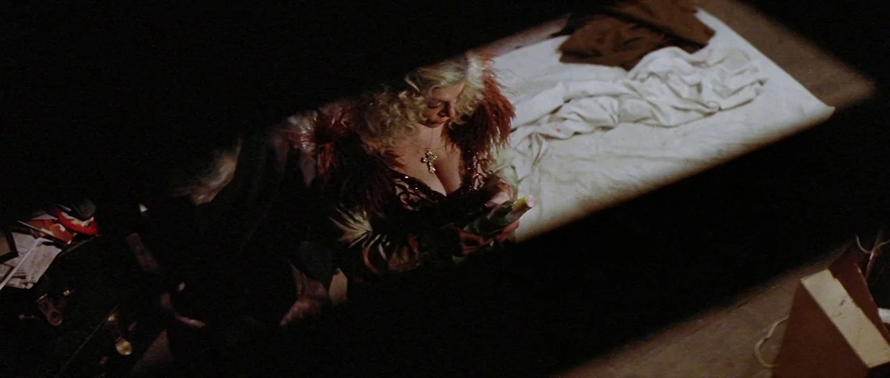 The Funhouse (1981) movie still: medium — A dark, cramped space contains a single bed with rumpled white sheets. A blonde woman in a lace garment…