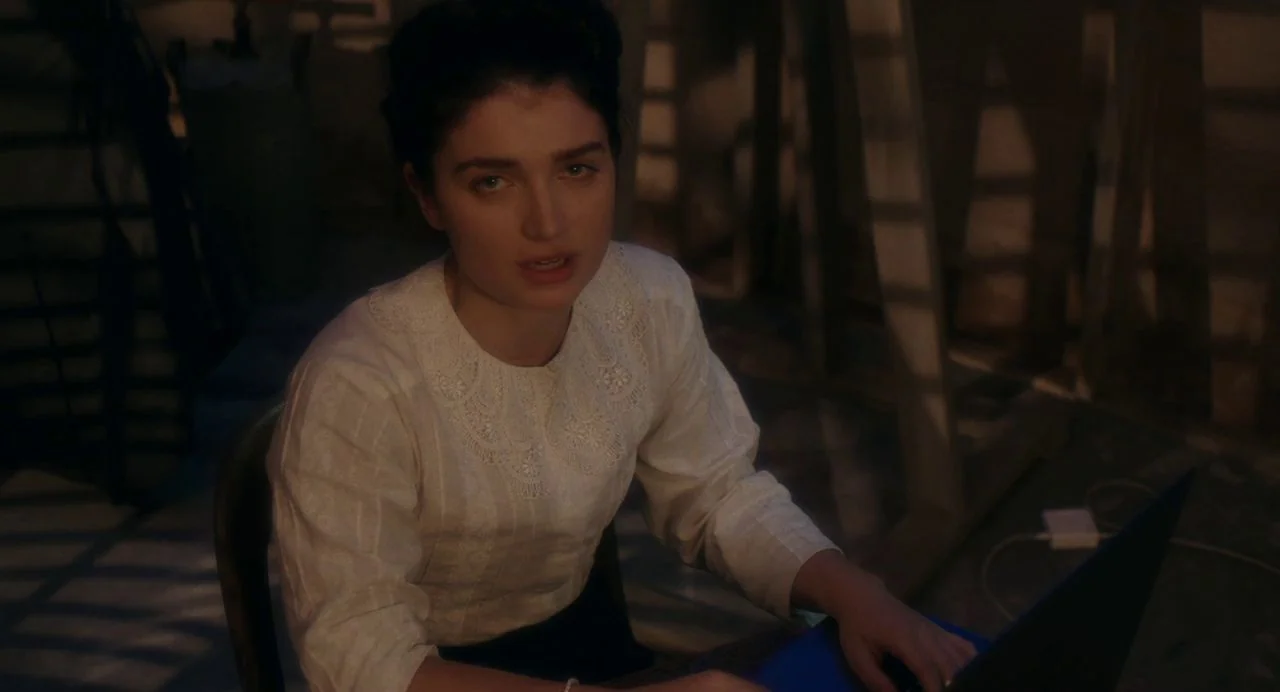 Tesla (2020) movie still: medium — A dimly lit attic room features wooden rafters casting long shadows across the space. A woman in a white…