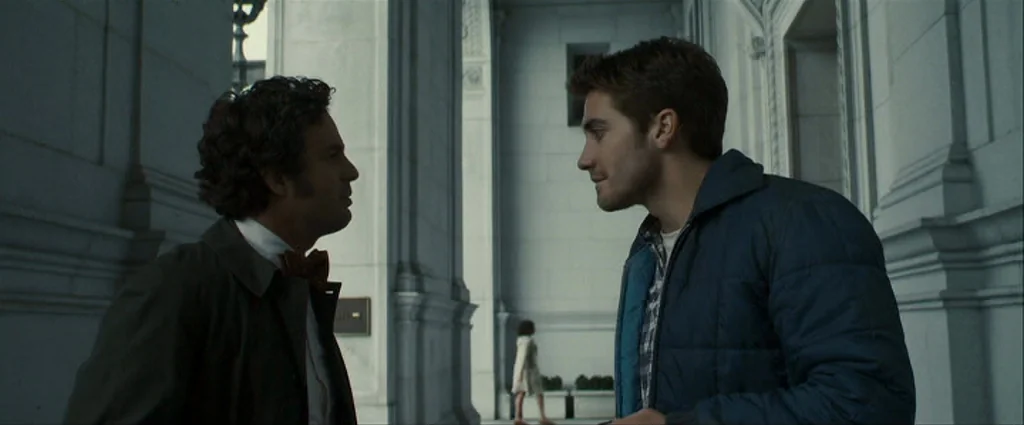 Zodiac (2007) movie still: medium — A stone colonnade with massive pillars defines the structural backdrop of the walkway. Two men stand facing each…