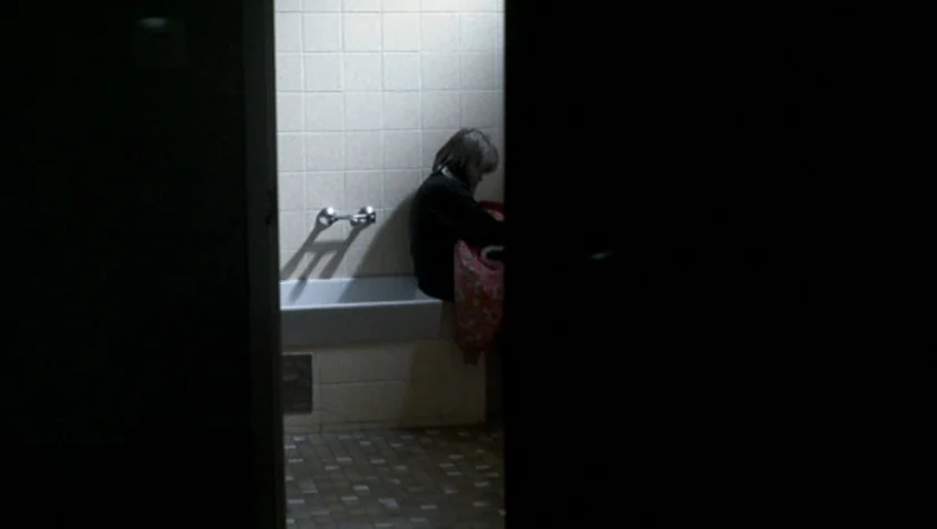 71 Fragments of a Chronology of Chance (1994) movie still: medium — A dimly lit room with light beige tiled walls and a basin fixture. A woman sits…