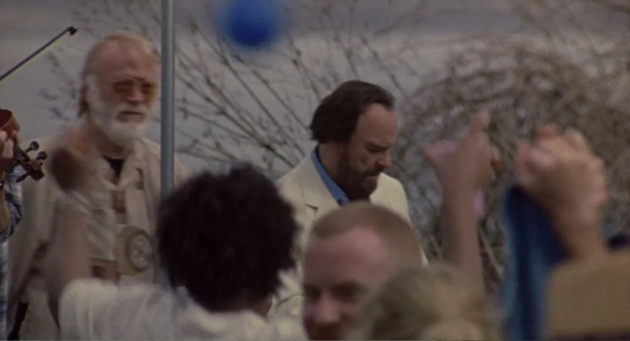 Forty Shades of Blue (2005) movie still: medium — A group of people gathers outdoors in a field with dry vegetation in background. A man plays the…