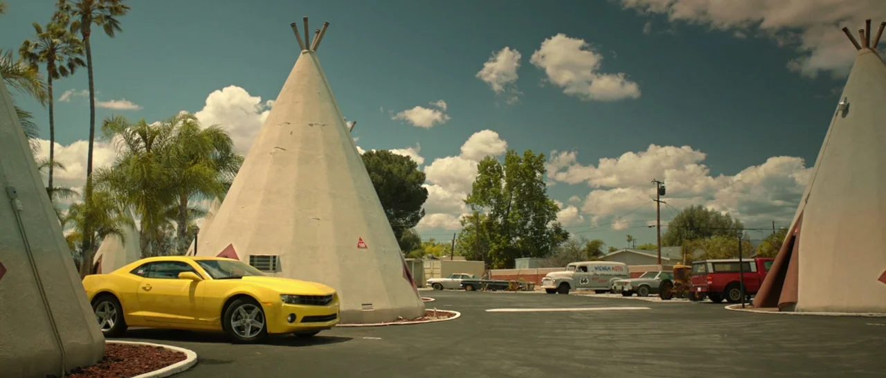 God Bless America (2011) movie still: wide — A sunny motel exterior features several white concrete teepee-shaped cabins arranged in a lot. A bright…
