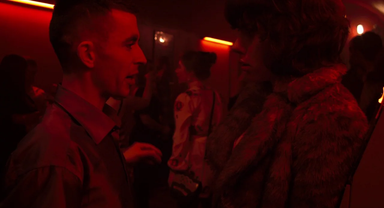 Under The Skin (2013) movie still: medium — A crowded dimly lit nightclub interior with red neon lights mounted on the ceiling. A man in a dark…