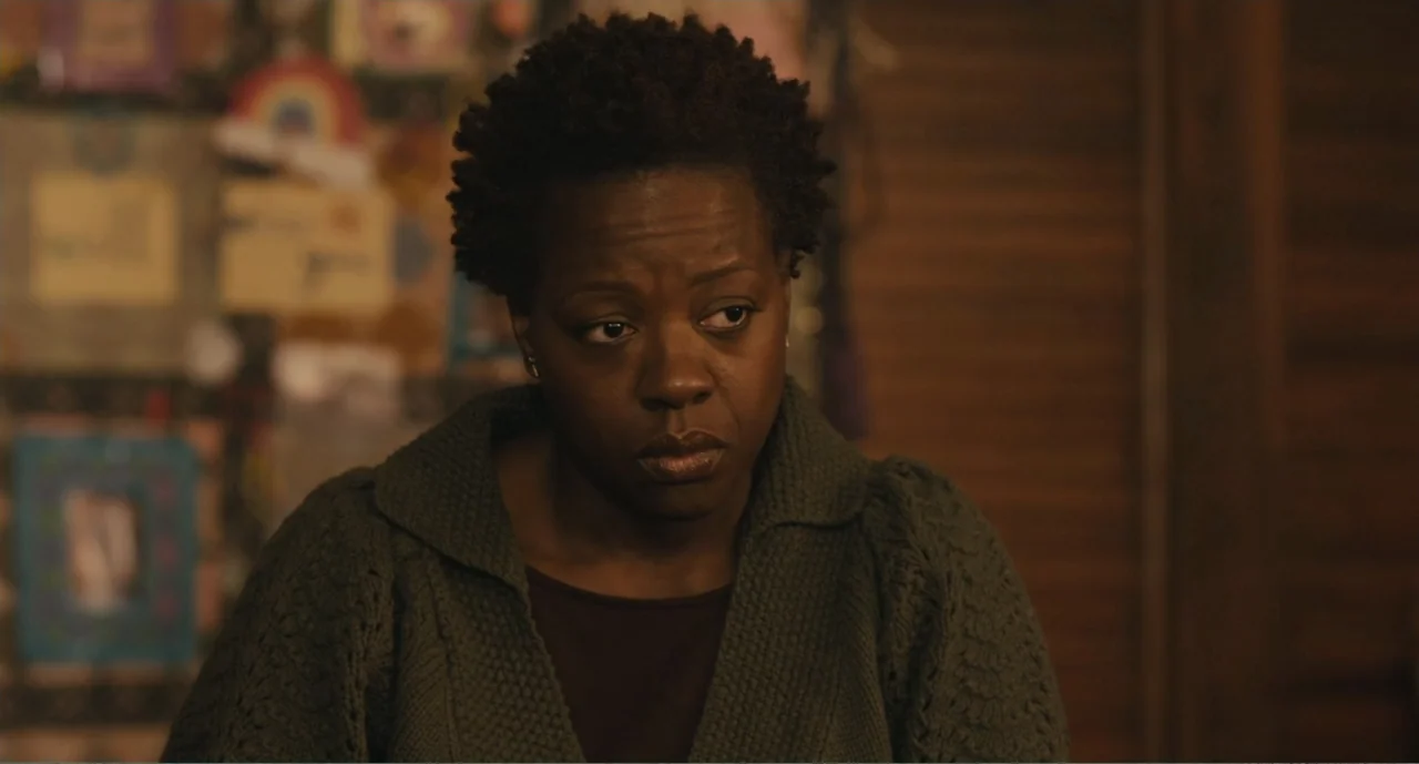 Lila & Eve (2015) movie still: medium — A dark indoor room with a wall decorated with multiple framed pictures. A woman with short textured hair…