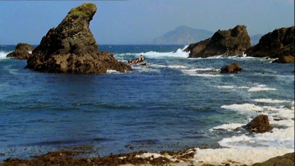 The Scarlet Letter (1973) movie still: wide — A rugged coastline features dark jagged rock formations jutting from the deep blue sea. A small…