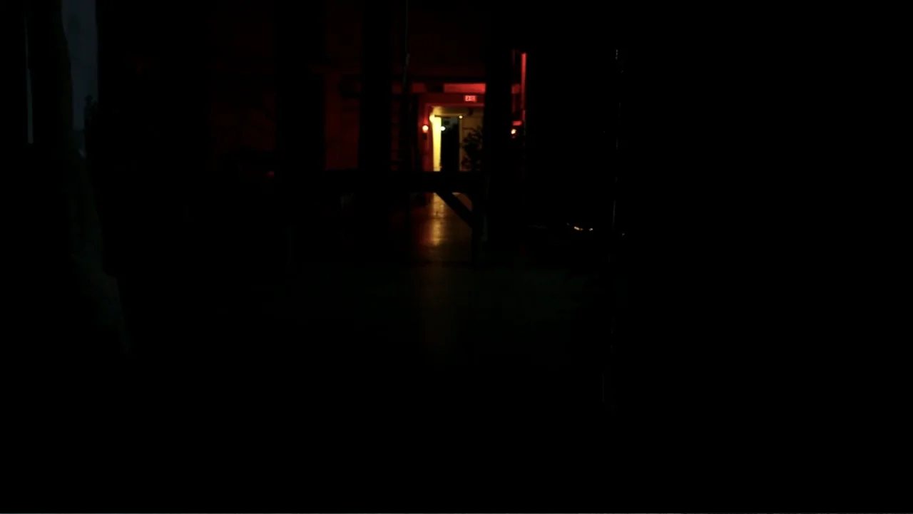 The Gallows (2015) movie still: establishing — A dark, empty industrial hallway terminates in a doorway marked with a red exit sign. No people are…