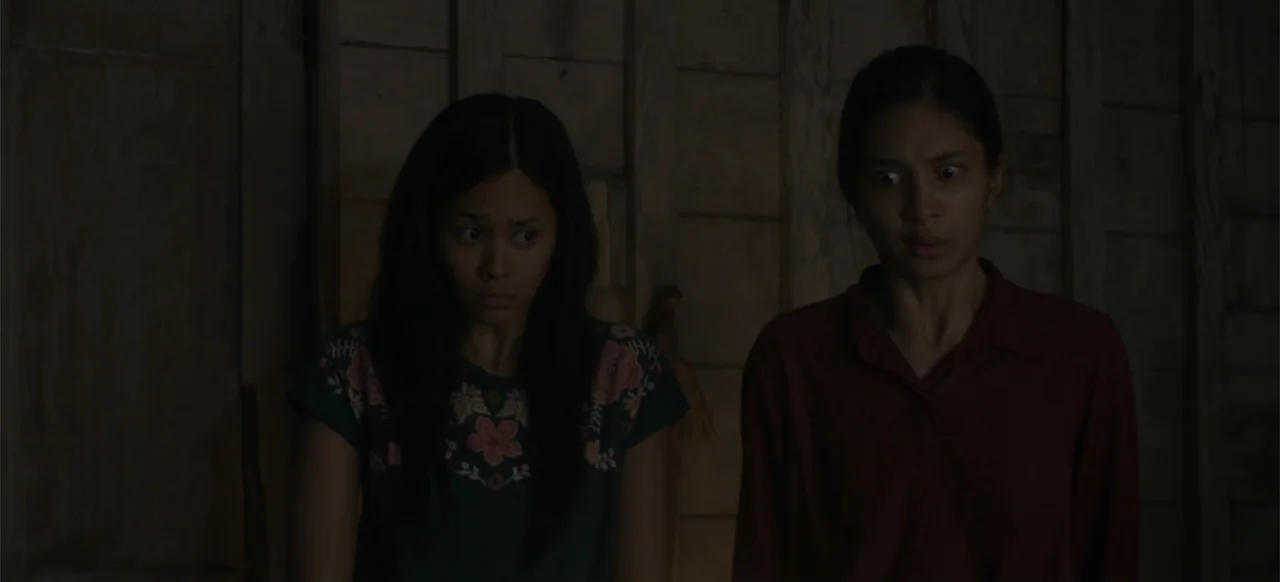 A Thousand Days (2023) movie still: medium — Two women stand in a dark, poorly lit room with vertical wooden plank walls. Both women gaze forward…