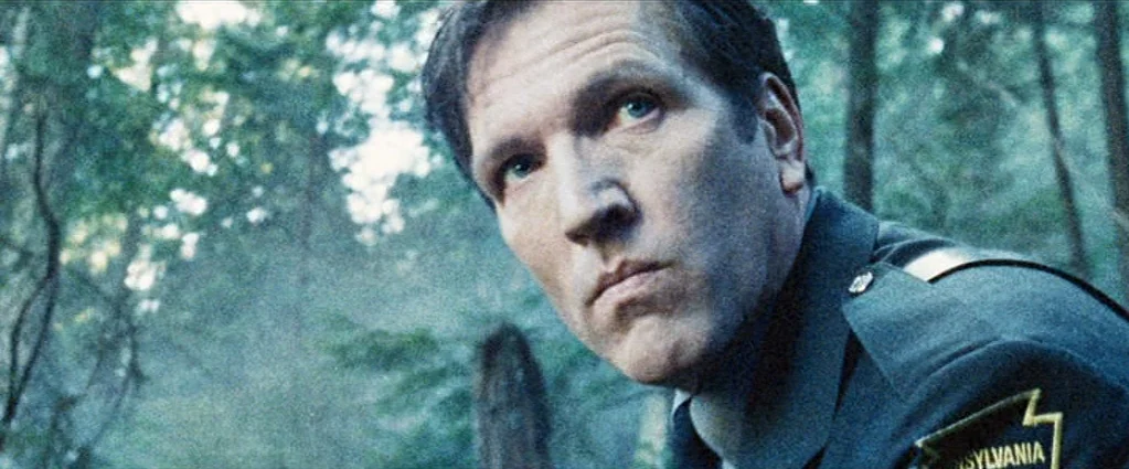Wind Chill (2007) movie still: closeup — A dense forest environment with blurred trees forming a natural background for the subject. A man in a dark…