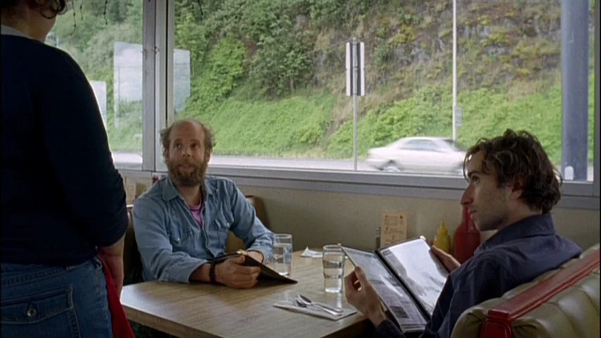 Old Joy (2006) movie still: medium — A diner booth faces a large window overlooking a sloped roadside with green vegetation. Two men sit at the…