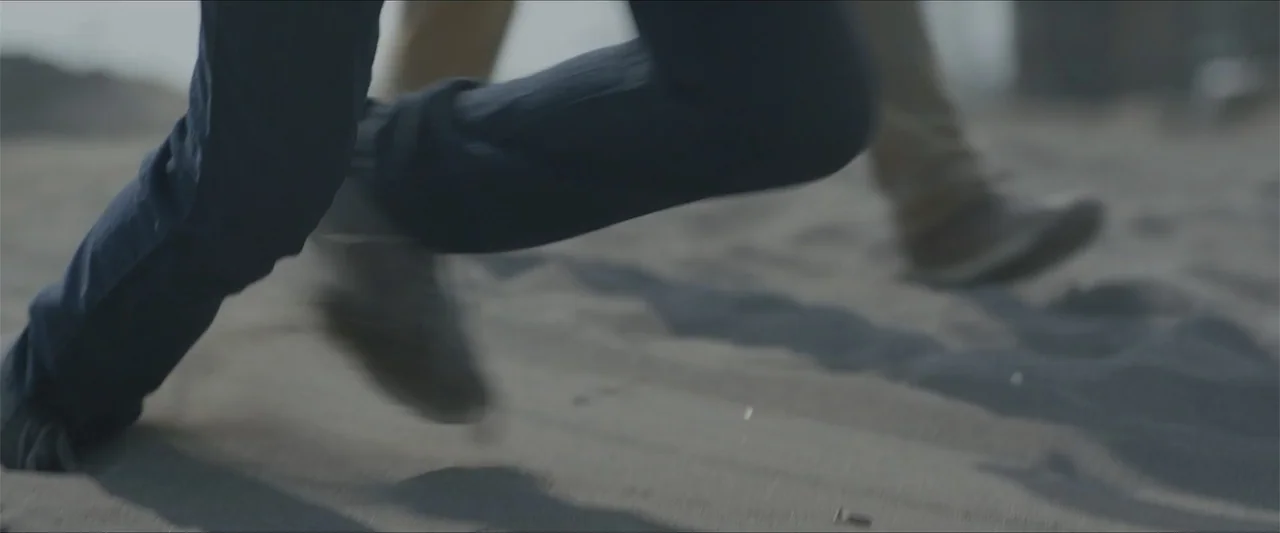 Radical (2023) movie still: closeup — A stretch of sandy ground is shown at ground level during the day. Feet of two individuals run quickly across…