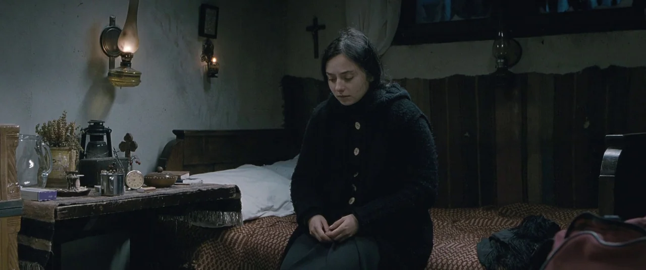 Beyond the Hills (2012) movie still: medium — A dimly lit, sparse room contains a wooden bed and a cluttered side table. A young woman sits silently…
