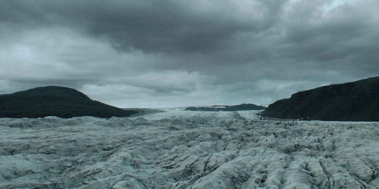 The Northman (2022) movie still: wide — A expansive glacier with deep crevices and uneven frozen terrain under heavy clouds. Dark rocky mountains…