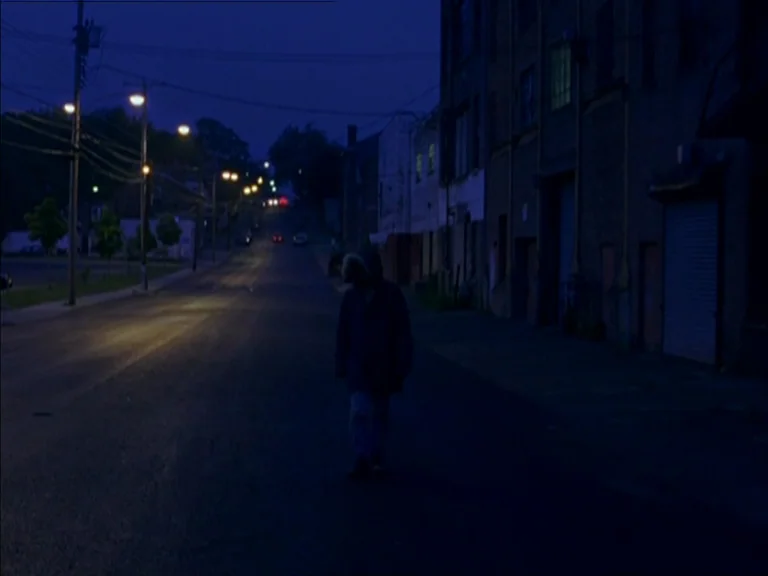 Last Days (2005) movie still: wide — A quiet urban street at night is lined with brick buildings and tall streetlamps. A lone hooded figure walks…