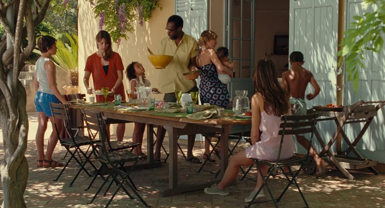 Young & Beautiful (2013) movie still: wide — A sunny stone-paved terrace features a large wooden dining table populated by a family. Adults pass…