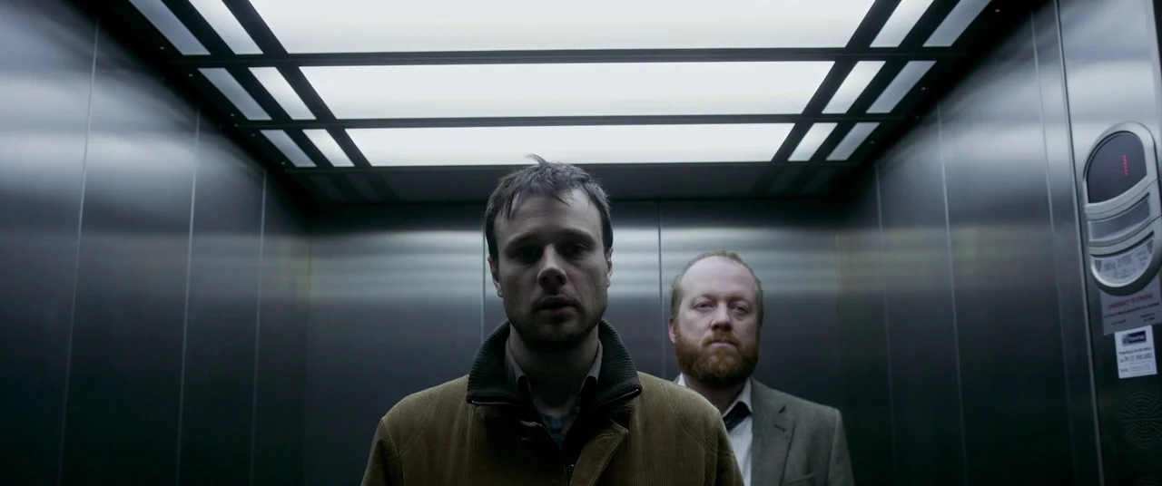 The Canal (2014) movie still: medium — Two men stand inside the metallic, clinical confines of a modern passenger elevator. The men are positioned…