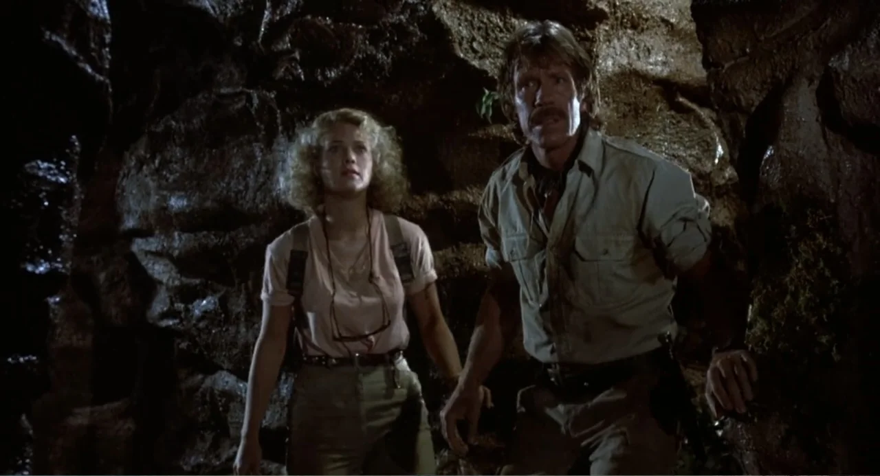 Firewalker (1986) movie still: medium — Two individuals stand inside a dark, jagged natural cave with damp rock walls. They look toward the upper…