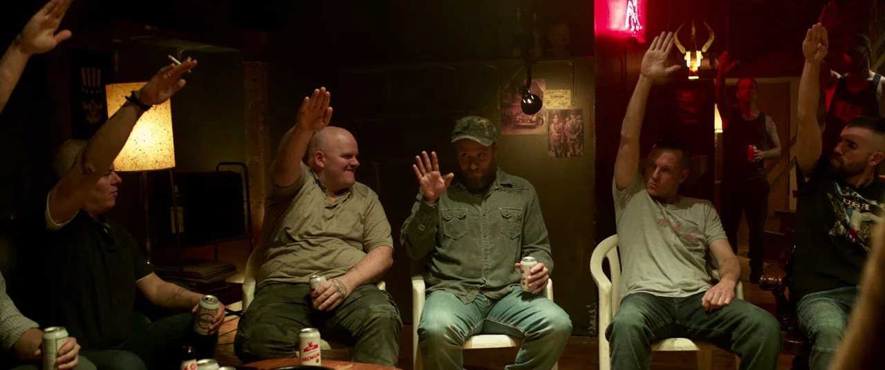 Long Shot (2019) movie still: wide — A dimly lit basement room with concrete walls features posters and a red neon sign. A group of men in casual…