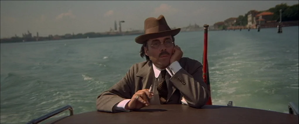 Death In Venice (1971) movie still: medium — A man rides in a small motorboat across a calm body of water. Distant buildings are visible on the…