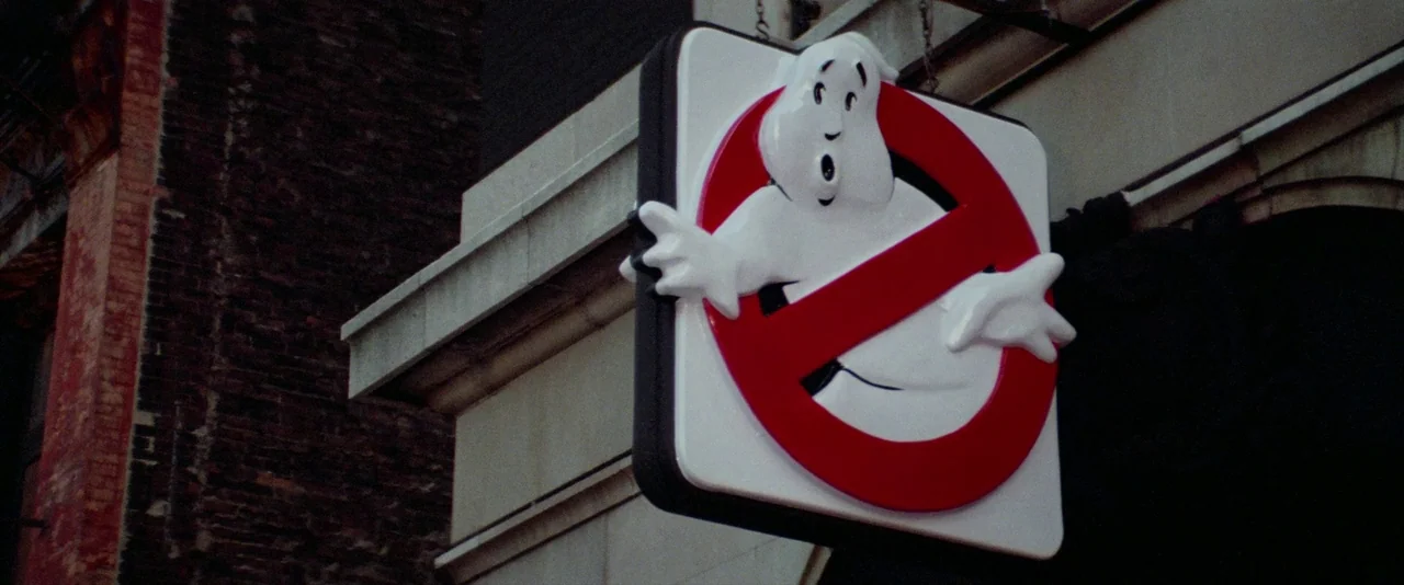 Ghostbusters (1984) movie still: closeup — A square Ghostbusters icon is mounted on the corner of a dark brick building. The sign features a…
