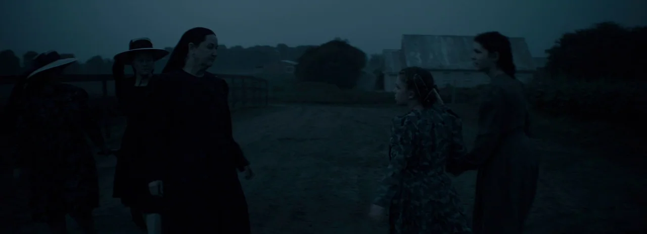 Women Talking (2022) movie still: wide — A dark rural field during the blue hour with an old wooden building behind. A group of four women dressed…