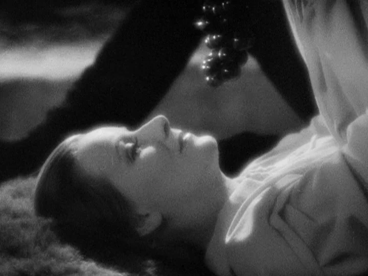 Queen Christina (1933) movie still: closeup — A dark, textured surface suggests a patch of grass in a studio setting. A woman lies on her back…