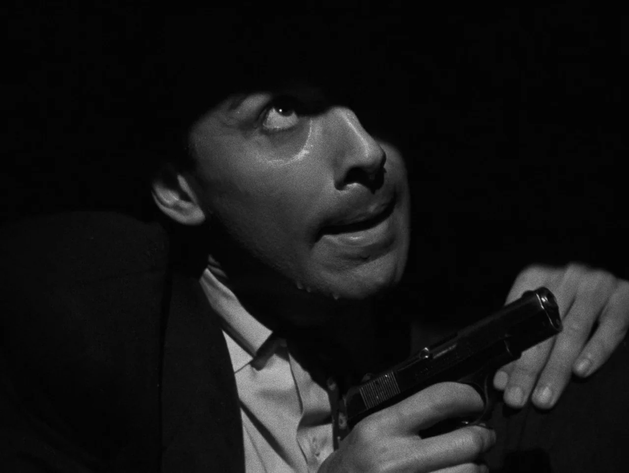 Pickup on South Street (1953) movie still: closeup — A dimly lit environment with no visible architectural details due to heavy shadow. A man with a…