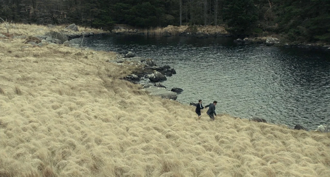 The Lobster (2015) movie still: wide — A wide shot shows a rocky shoreline adjacent to a dark, calm lake. A man and a woman in business attire run…
