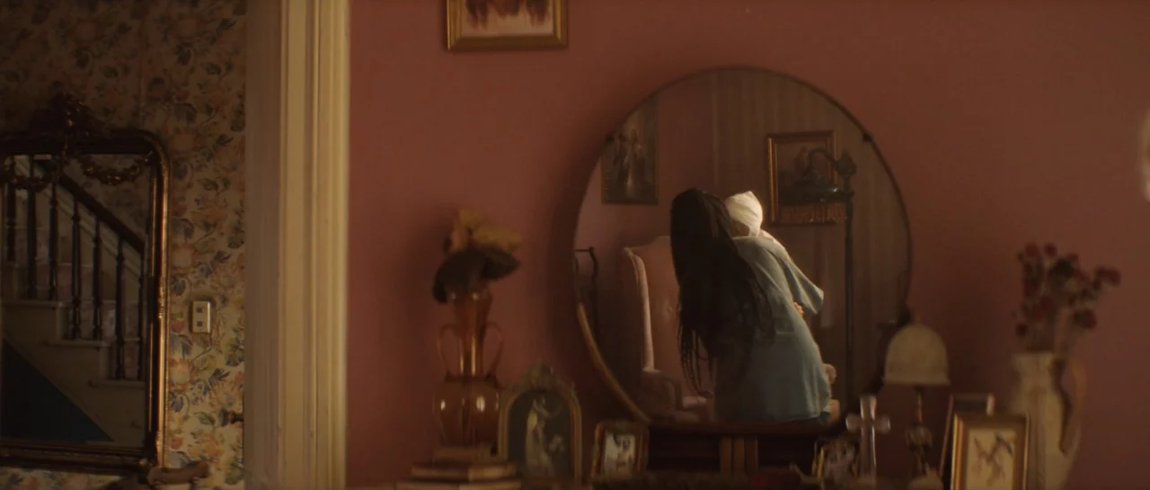 The Front Room (2024) movie still: medium — A room with pink walls and floral wallpaper contains a vanity and mirror. A person with dark braided…