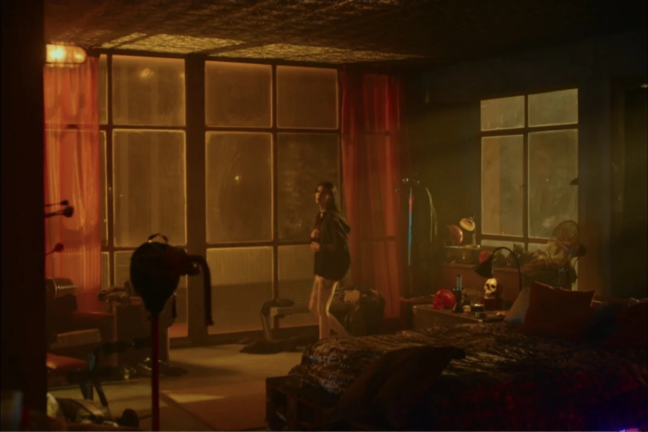 Cross the Line (2020) movie still: fullbody — A cluttered apartment bedroom features large window panes with thin red curtains. A young woman stands…