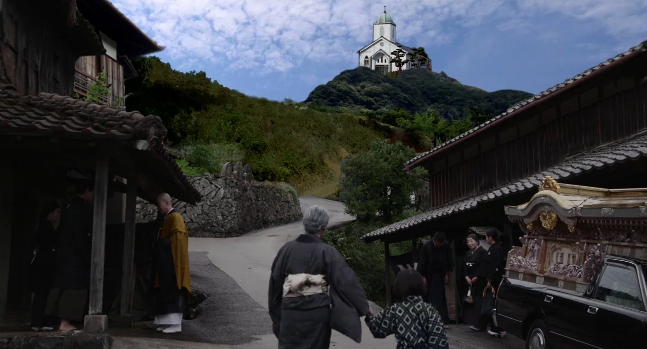 Hanagatami movie still: wide — A rural Japanese hillside village features traditional wooden houses and a winding paved path. A funeral hearse is…