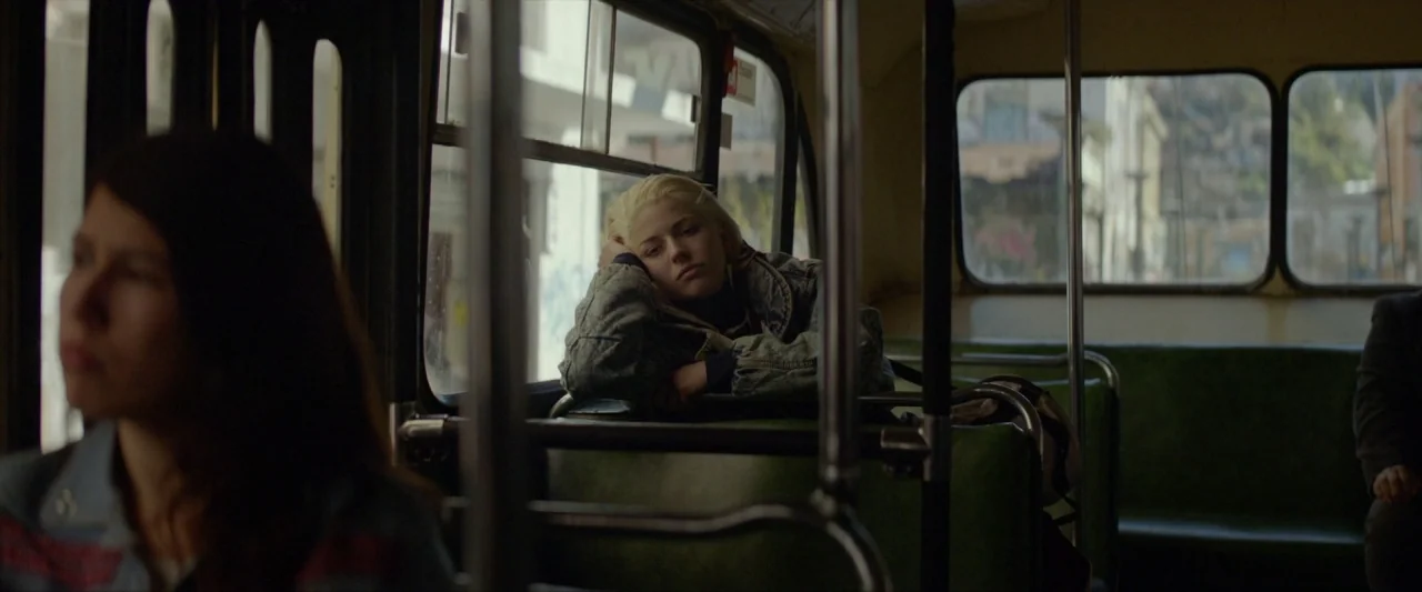 Ema (2019) movie still: medium — The interior of a bus with green vinyl seats and metal handrails. A woman with short blonde hair sits by the window…