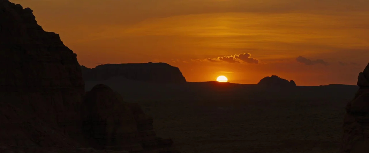 Damsel (2018) movie still: establishing — A vast arid desert landscape features towering jagged rock formations under an orange sky. The sun dips…