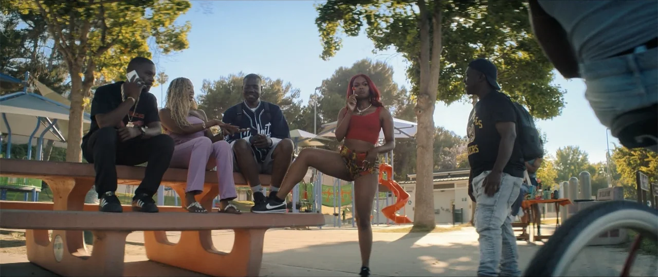One of Them Days (2025) movie still: fullbody — A sunny public park features mature green trees and playground structures in the background. A group…