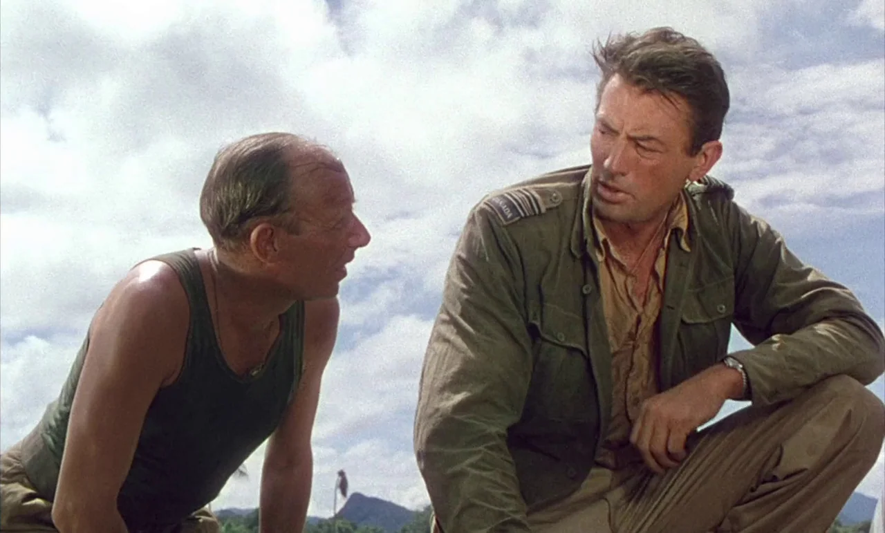 The Purple Plain (1954) movie still: medium — Two men in military clothing kneel on a grassy landscape under a cloudy sky. The man on the right…