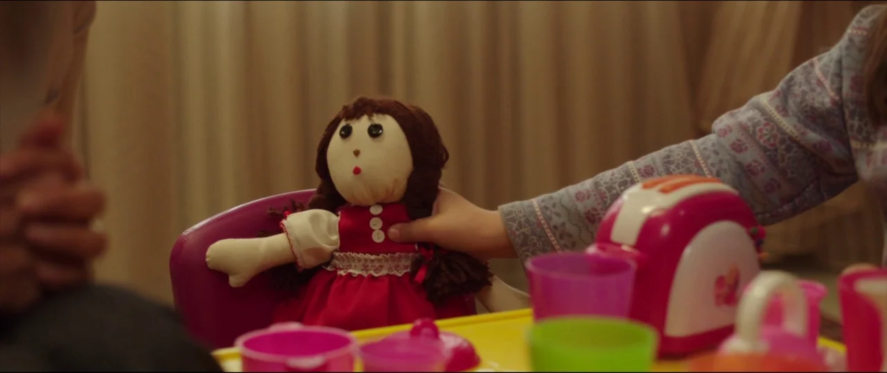 Under The Shadow (2016) movie still: medium — A toy rag doll in a red dress sits on a small pink chair. A child's hand reaches out to touch the…