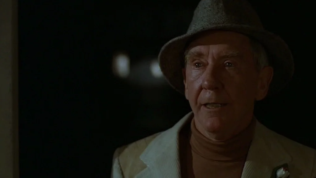 Magic (1978) movie still: closeup — An older man stands in a poorly lit space against a dark, featureless backdrop. The man is wearing a hat and…