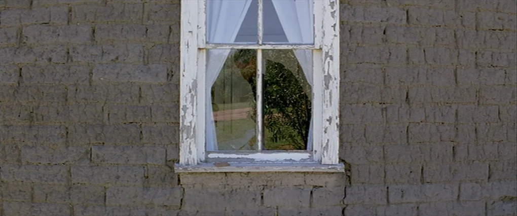 Silent Light (2007) movie still: medium — An exterior wall made of cracked adobe blocks features a single weathered white window. A man is visible…