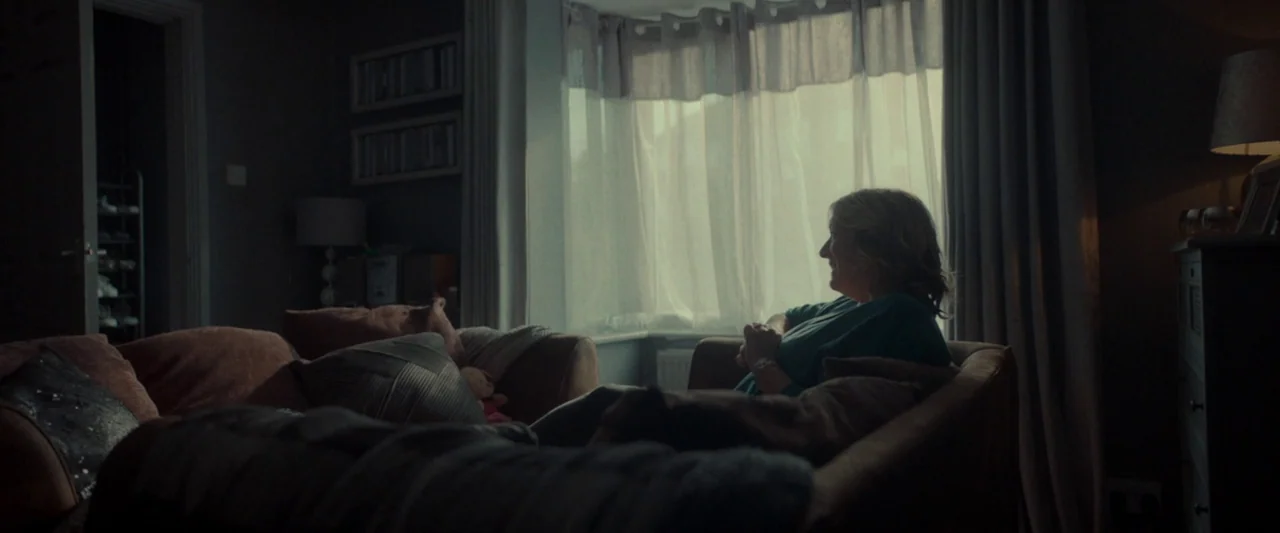 Ali & Ava (2021) movie still: medium — A dimly lit room features a sofa facing a bright window with sheer curtains. A woman sits on the sofa and…