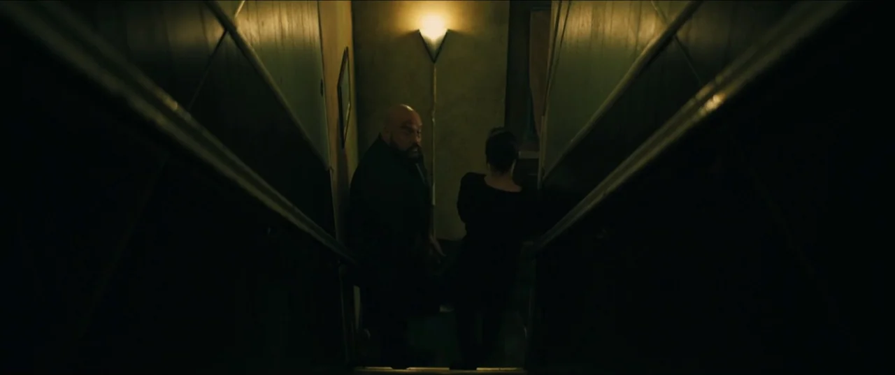 Guns Up (2025) movie still: medium — A dark, narrow stairwell with muted green walls and dimly lit wooden banisters. A bearded man and a woman in a…