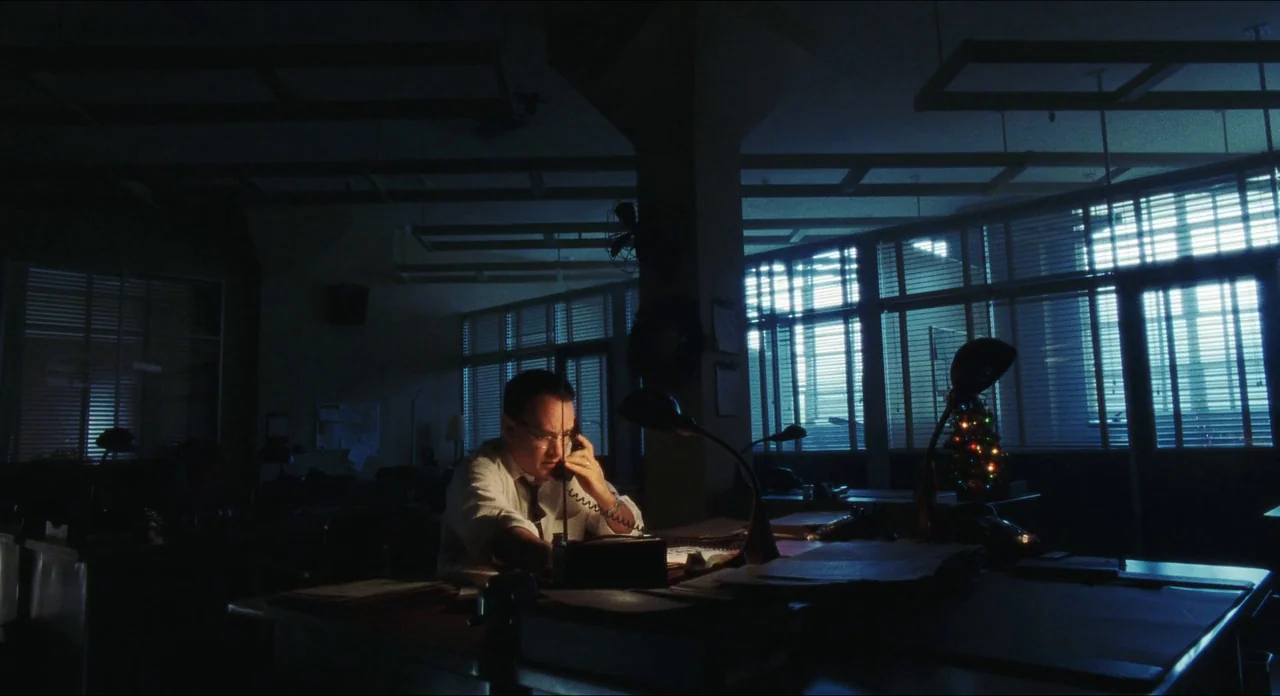 Catch Me If You Can (2002) movie still: medium — An open plan workspace at night is lit by a single desk lamp. A man sits alone at his desk speaking…