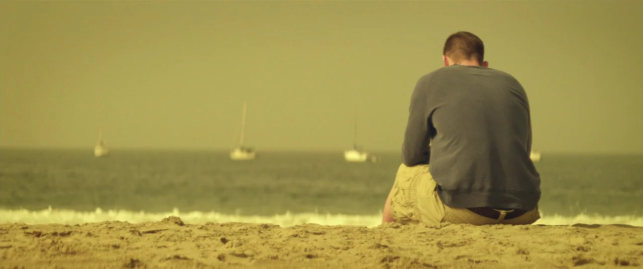 Magic Mike (2012) movie still: fullbody — A man sits on a sandy beach looking out toward the ocean. Several white sailboats are visible on the…