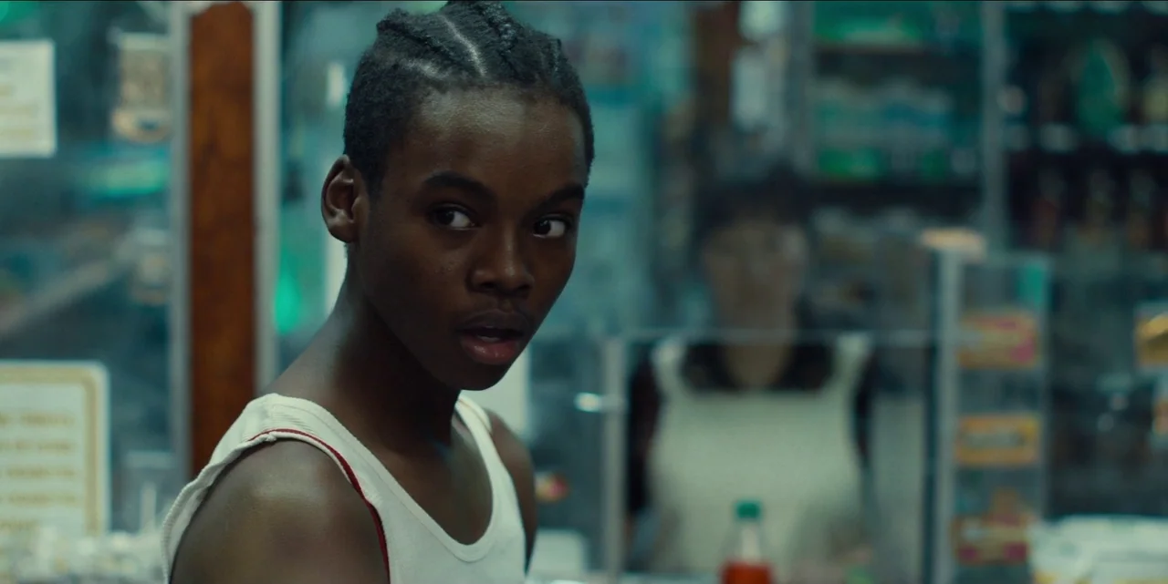 Charm City Kings (2020) movie still: medium — A dimly lit retail space features shelves stocked with various products in the background. A young…