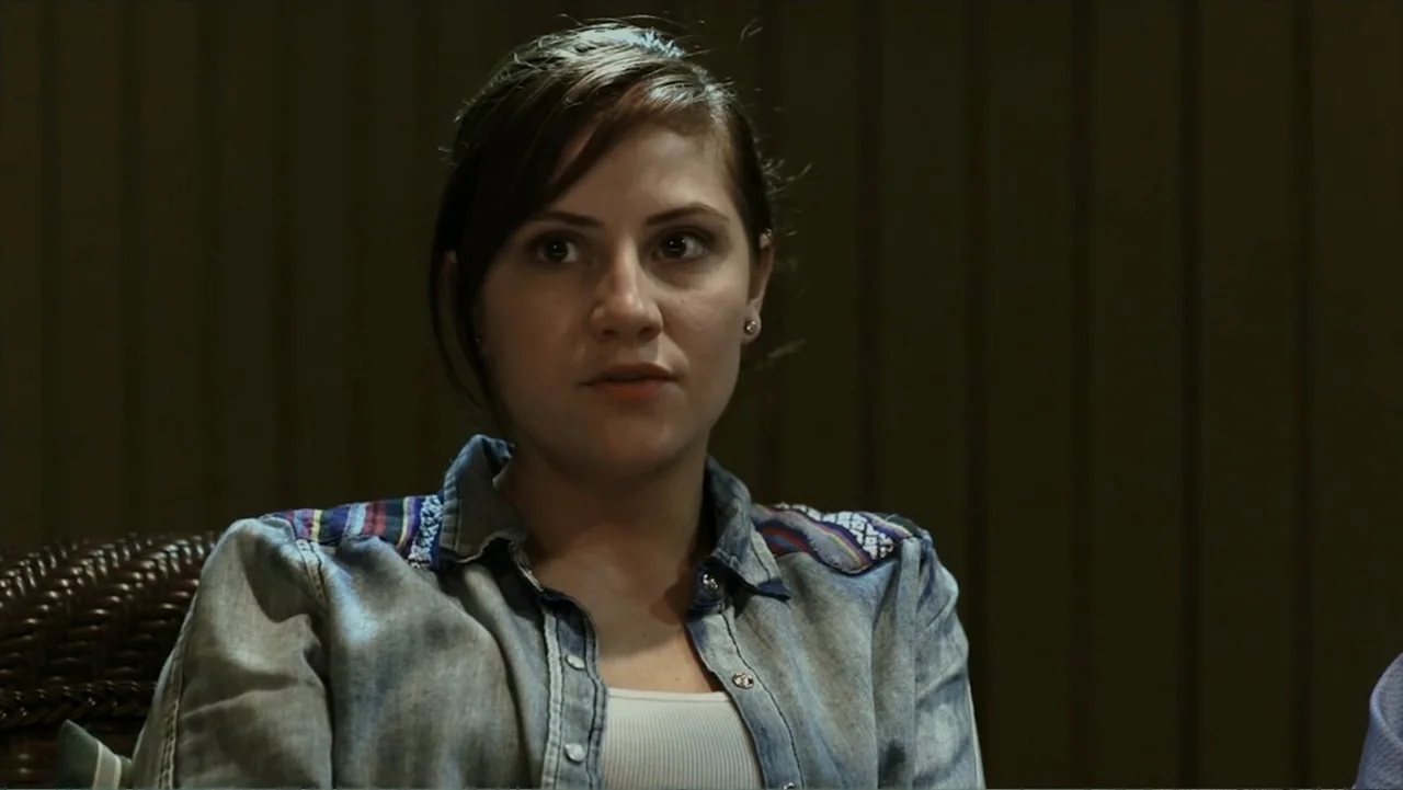 Hello I Love You (2018) movie still: medium — A dimly lit room with dark wooden wall paneling in the background. A woman in a denim shirt sits…