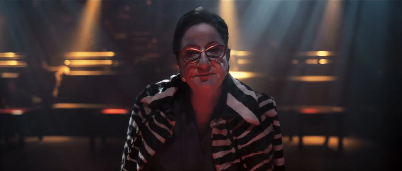 Historias lamentables (2021) movie still: medium — A dimly lit nightclub features rows of empty tables and overhead stage lighting beams. An older…