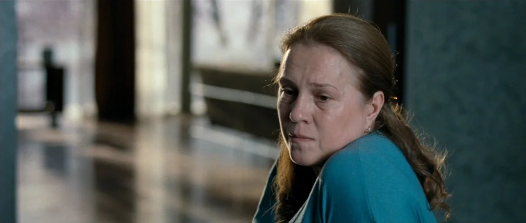 Elena (2011) movie still: medium — A living area with muted tones and a blurred background featuring a large window. A middle-aged woman is…