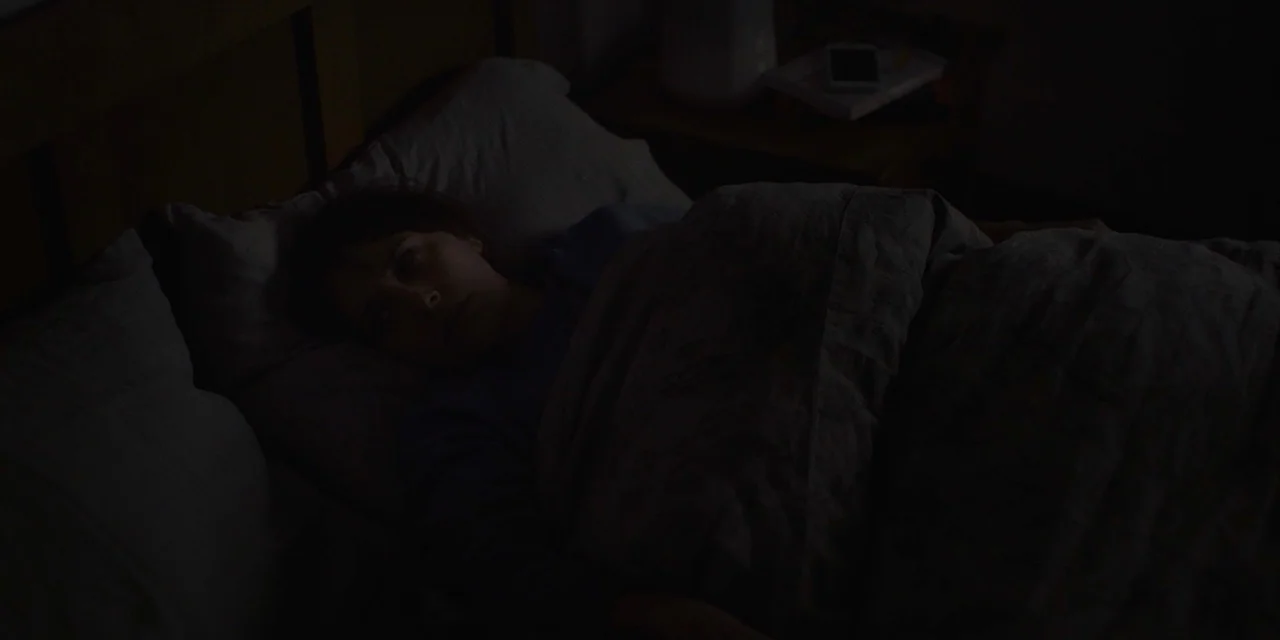 The Sound of Silence movie still: closeup — A person lies awake in a dark room under a gray blanket. The scene is illuminated only by a faint, cool…